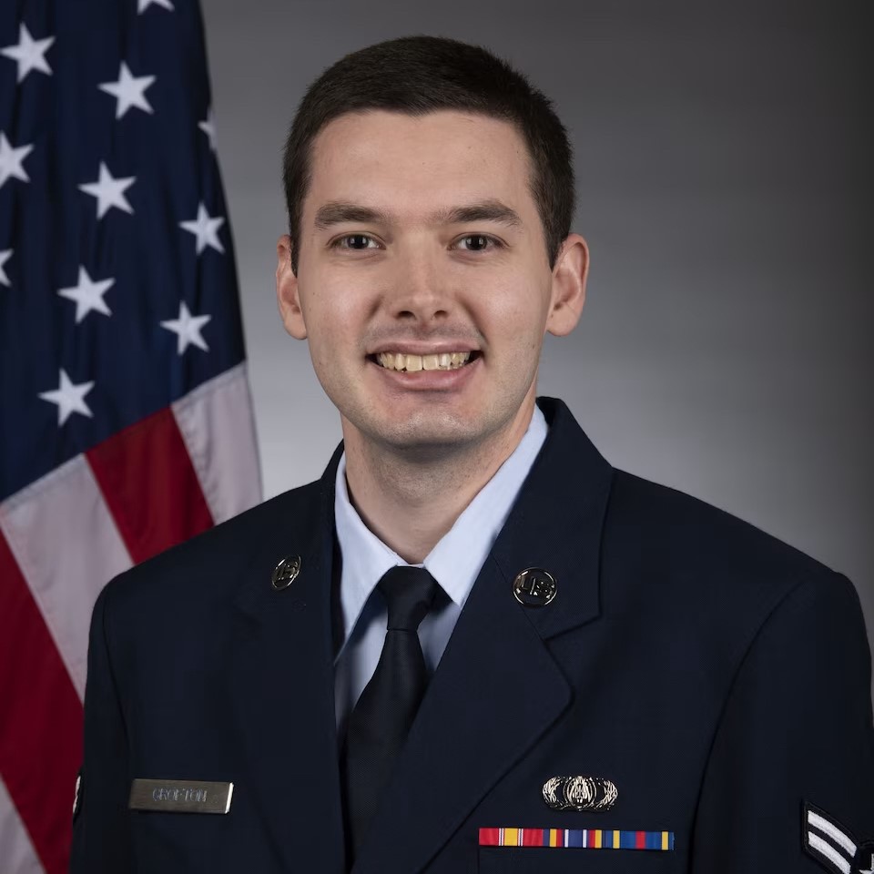 Man in a navy suit from the chest up, standing in front of an American flag
